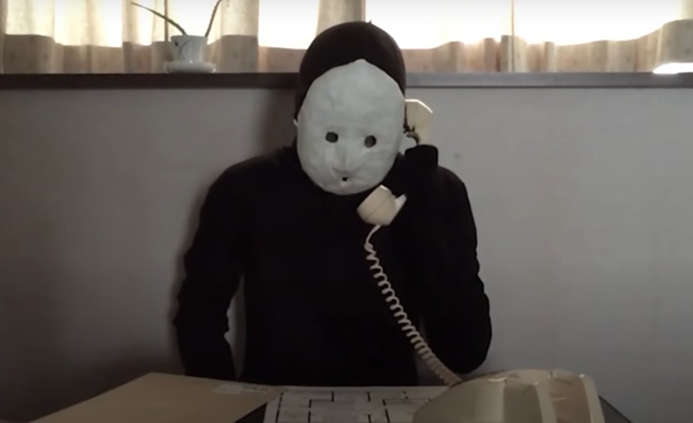 Uketsu, l'autore di Strani disegni, in tuta nera e maschera di cartapesta. Sta telefonando con un classico telefono a cornetta, e sul tavolo davanti a lui c'è la planimetria di una casa.